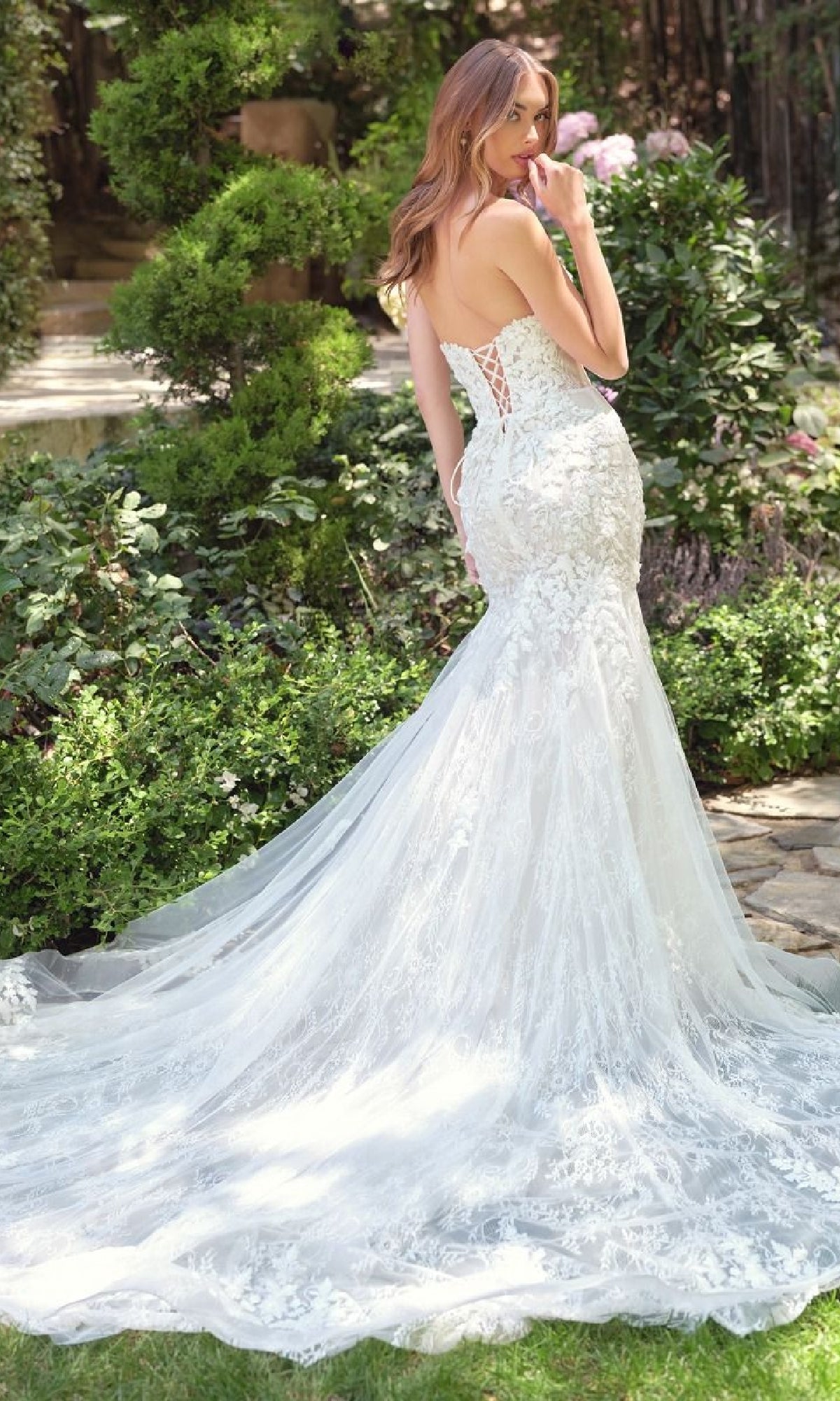 A woman stands in a garden, wearing the Andrea & Leo WL088 strapless mermaid bridal dress with a lace-up back, flowing train, and delicate floral lace details for a romantic touch.